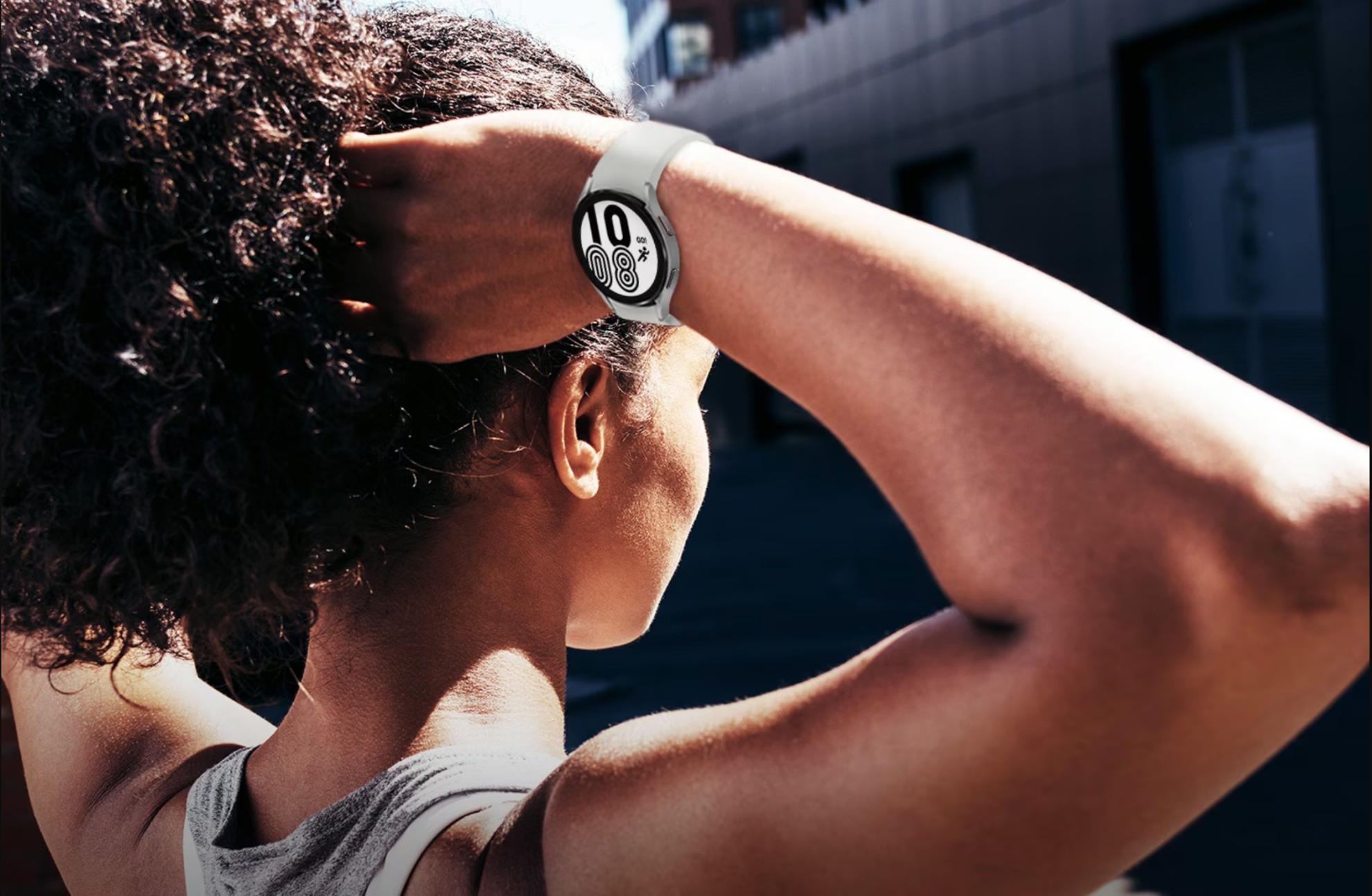 There is a woman outside wearing workout clothes and raising her hands to tie her hair. She is wearing a Galaxy Watch4 with a Silver color sport band. The watch is displaying the time and a UI icon with a running symbol that says †GO!' above.